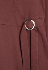 Szczegół burgundowej sukienki z gładkiego materiału, ze srebrnym akcentem w postaci pierścienia i wiązaną tasiemką w pasie, podkreślający strukturalny, minimalistyczny design.