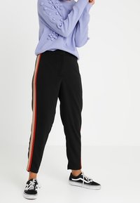 Osoba w czarnych spodniach z czerwonymi i beżowymi paskami po bokach, w fakturowanym lawendowym swetrze oraz czarno-białych trampkach, stoi z uniesionymi ramionami.