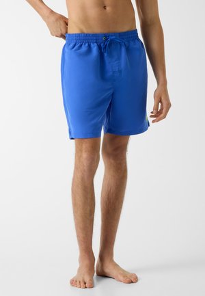Mann trägt blaue Badeshorts mit Kordelzug in der Taille, steht barfuß vor weißem Hintergrund.