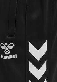 Svarta träningsbyxor med texturerad yta, med en vit Hummel-logotyp och vita chevron-mönster längs sidan.
