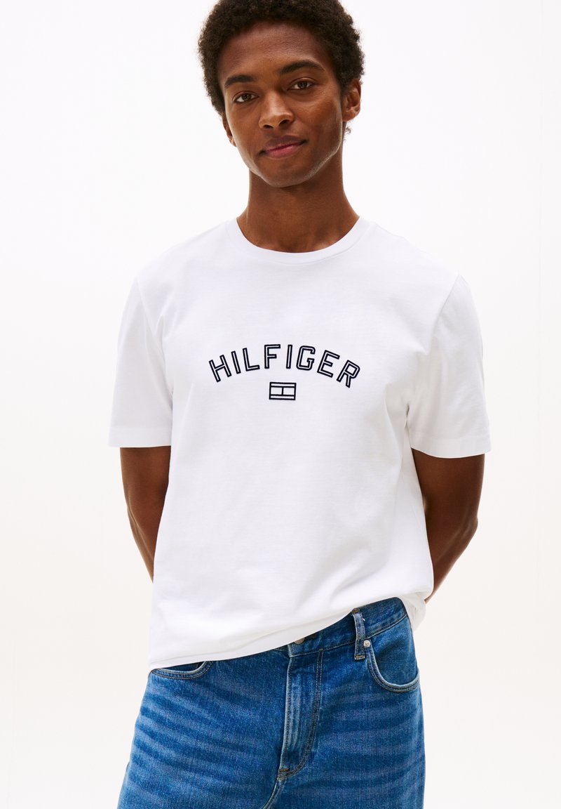 Witte katoenen t-shirt met een ronde hals en korte mouwen, voorzien van "HILFIGER" geborduurd in marineblauw over de borst. Draag het met blauwe jeans.
