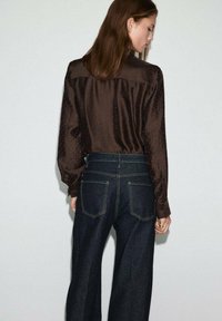 Blusa de satén marrón con textura de lunares, con mangas largas y un corte relajado, combinada con jeans de mezclilla oscura de cintura alta con bolsillos.