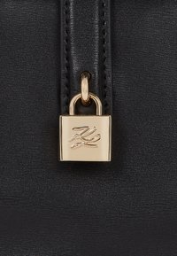 Λεπτομέρεια μαύρης δερμάτινης τσάντας με χρυσό λουκέτο και ανάγλυφο λογότυπο. Είναι ορατή η λεία υφή και η προσεγμένη ραφή.