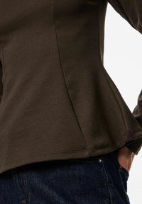 Brun långärmad topp med figurnära kropp och utsvängd nederdel, tillverkad av ett mjukt tyg. Synliga sömdetaljer och mörka denimjeans vid midjan.