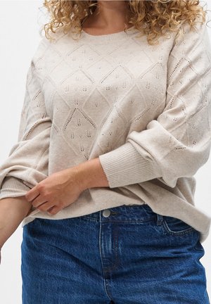 Femme portant un pull en maille beige avec des motifs en losange et un jean bleu en denim, un bras ajustant le poignet du pull.