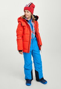 Veste matelassée rouge avec une capuche, chemise bleue à motifs, pantalon bleu et bottes foncées. Le design présente des couleurs et des textures contrastées.