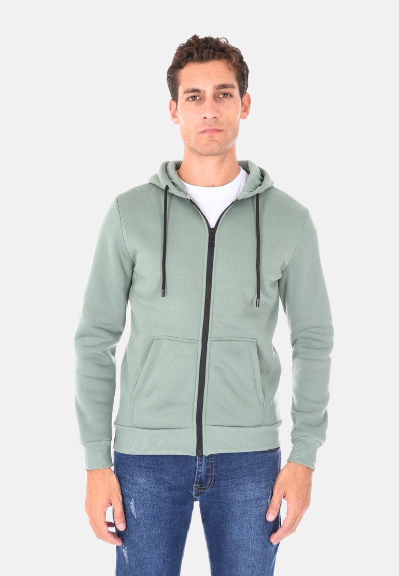 Felpa verde con zip, con lacci neri, tasche a marsupio, polsini a costine e una texture di tessuto morbida. Indossata sopra una maglietta bianca.