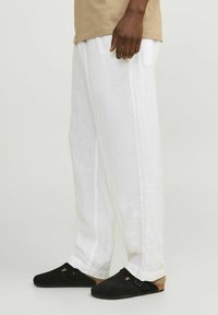 Osoba ubrana w luźne białe lniane spodnie i czarne klapki na płaskiej podeszwie z korka, stojąca na jednolitym tle.