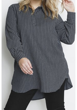 Femme portant une chemise à carreaux noir et blanc ample à manches longues avec un pantalon noir, posant avec une main près de ses cheveux.