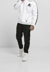 Uomo che indossa un hoodie bianco della Starter con logo nero, pantaloni cargo neri e sneaker alte bianche mentre si trova su uno sfondo grigio.