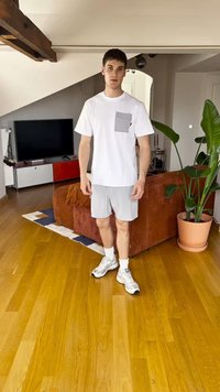 Bílé tričko s krátkým rukávem a šedou kapsou, šedé šortky, bílé ponožky a šedé tenisky. Stojí na dřevěné podlaze v místnosti s rostlinami.