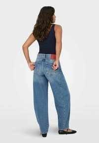 Jeans in denim blu a vita alta e gamba larga con una finitura sbiadita e un'etichetta in pelle rossa sulla parte posteriore della vita. Modella che indossa scarpe basse.