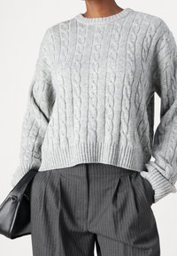 Pull gris en maille torsadée avec col rond et ourlet côtelé, associé à un pantalon gris à fines rayures et une pochette en cuir noire.