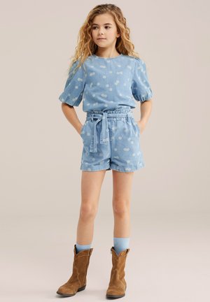 Mlada deklica z kodrastimi lasmi, oblečena v komplet modre obleke z rožnatim vzorcem in kratke hlače s pasom, svetlo modre nogavice in rjave antilop kavbojske škornje.