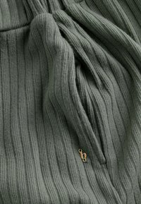 Groene ribbelstof met een zachte textuur, voorzien van een klein gouden accentdetail. Het materiaal lijkt licht en vloeiend.