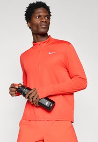 Long-sleeved, zippered athletic top in bright red with a gray Nike logo. Model holds a black water bottle with a sleek design.