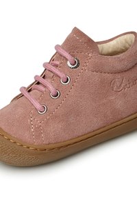 Zapato de ante para bebé en rosa suave con cordones a juego. Cuenta con ojales metálicos, una punta redondeada y una suela de goma texturizada con un borde marrón.