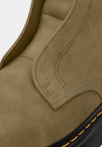 Primo piano di un stivale in suede beige con cuciture dettagliate e suola nera con accenti gialli nella cucitura.