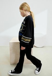 Svart sweatshirt med leopardmönstrade detaljer, långa ärmar och grafisk design, ihop med svarta ribbade byxor och svarta sneakers med vita sulor.