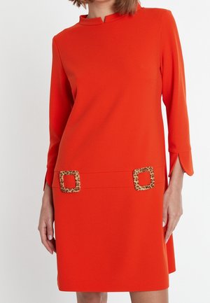 Vrouw draagt een feloranje jurk met lange mouwen, een gespleten halslijn en twee vierkante riemaccenten met luipaardprint op de heupen.