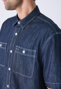 Camisa corta de manga azul oscura en tela de mezclilla, con costuras blancas, dos bolsillos en el pecho y un cuello clásico con cierres de botón.