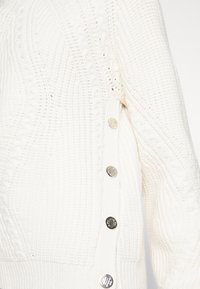 Manche de pull en maille couleur écru avec motifs texturés et quatre boutons métalliques le long de la couture latérale.