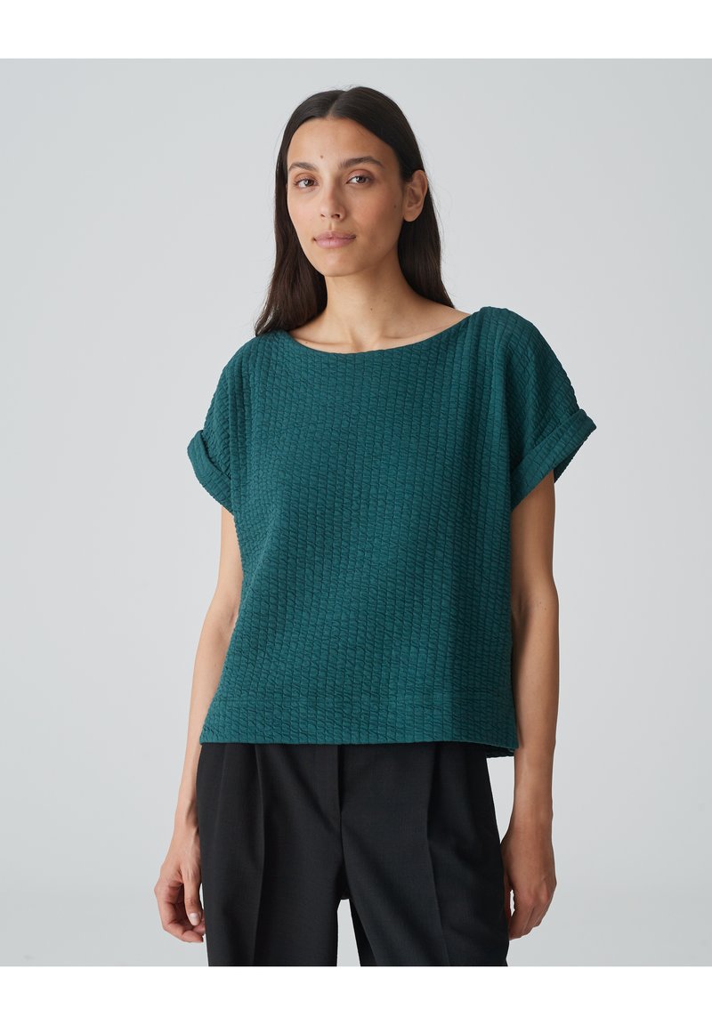 Teal textuur short-sleeve top met een brede halslijn en omgeslagen mouwen, gecombineerd met losse zwarte broeken. Eenvoudig ontwerp met een relaxte pasvorm.