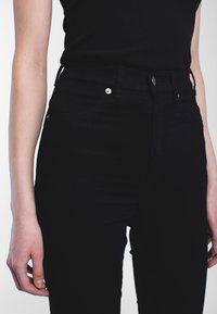 Jeans neri a vita alta in denim, con tasche anteriori, zip e accessori in metallo. Il tessuto ha una consistenza liscia.