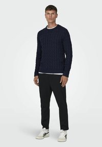 Marineblauwe kabelgebreide trui met geribbelde manchetten en zoom, gedragen over een wit T-shirt met ronde hals, in combinatie met zwarte broek en witte sneakers.