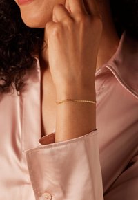 Gold chain bracelet on wrist, featuring small circular links. Worn with a light pink satin shirt, showing smooth texture and elegant design.