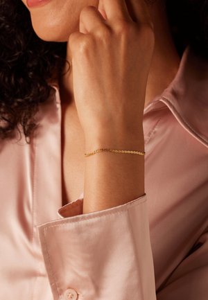 Gouden schakelsub bracelet om de pols, met kleine ronde schakken. Draagt een lichtroze satijnen shirt, dat een gladde textuur en elegant ontwerp toont.