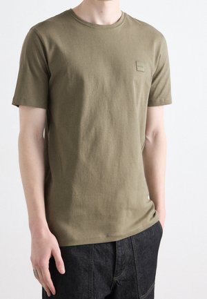 Person trägt ein schlichtes olivgrünes T-Shirt mit kurzen Ärmeln, einem kleinen quadratischen Logo auf der Brust und dunklen Hosen vor weißem Hintergrund.