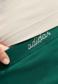 Krémový vrch s vrstvou nad tmavozelenými šortkami, ktoré majú textúrovanú tkaninu a vyšívané logo "adidas" na páse.