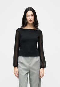 Γυναίκα με μακριά σκούρα μαλλιά φορώντας μαύρο διαφανές μακρυμάνικο τοπ και ανοιχτό γκρι παντελόνι με λεπτές ρίγες, όρθια μπροστά σε απλό λευκό φόντο.