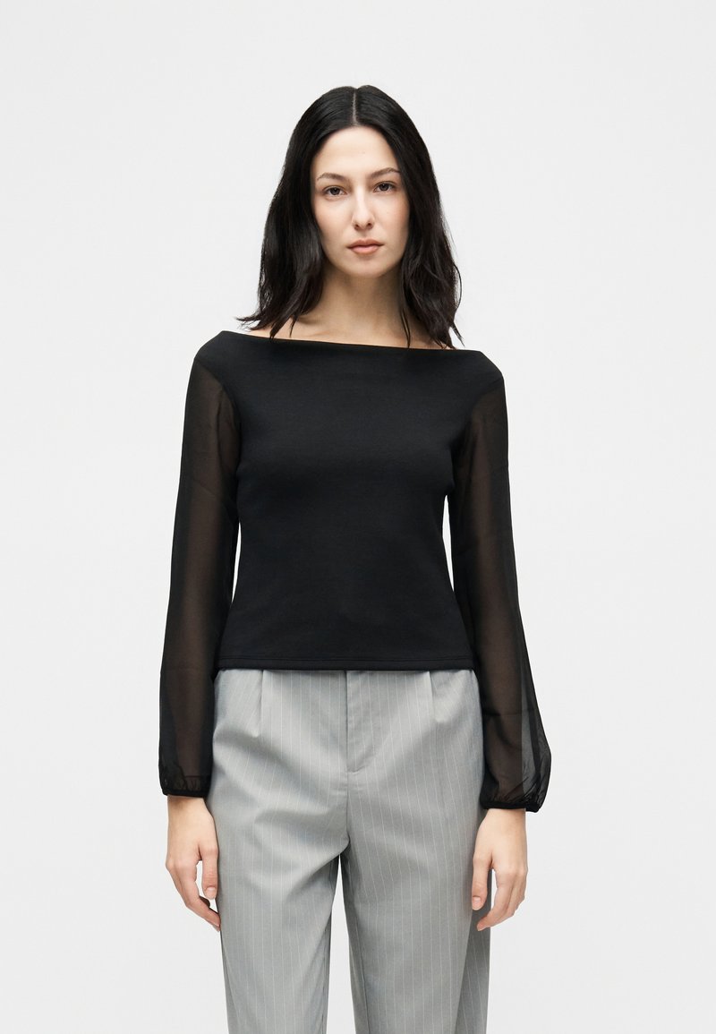 Γυναίκα με μακριά σκούρα μαλλιά φορώντας μαύρο διαφανές μακρυμάνικο τοπ και ανοιχτό γκρι παντελόνι με λεπτές ρίγες, όρθια μπροστά σε απλό λευκό φόντο.