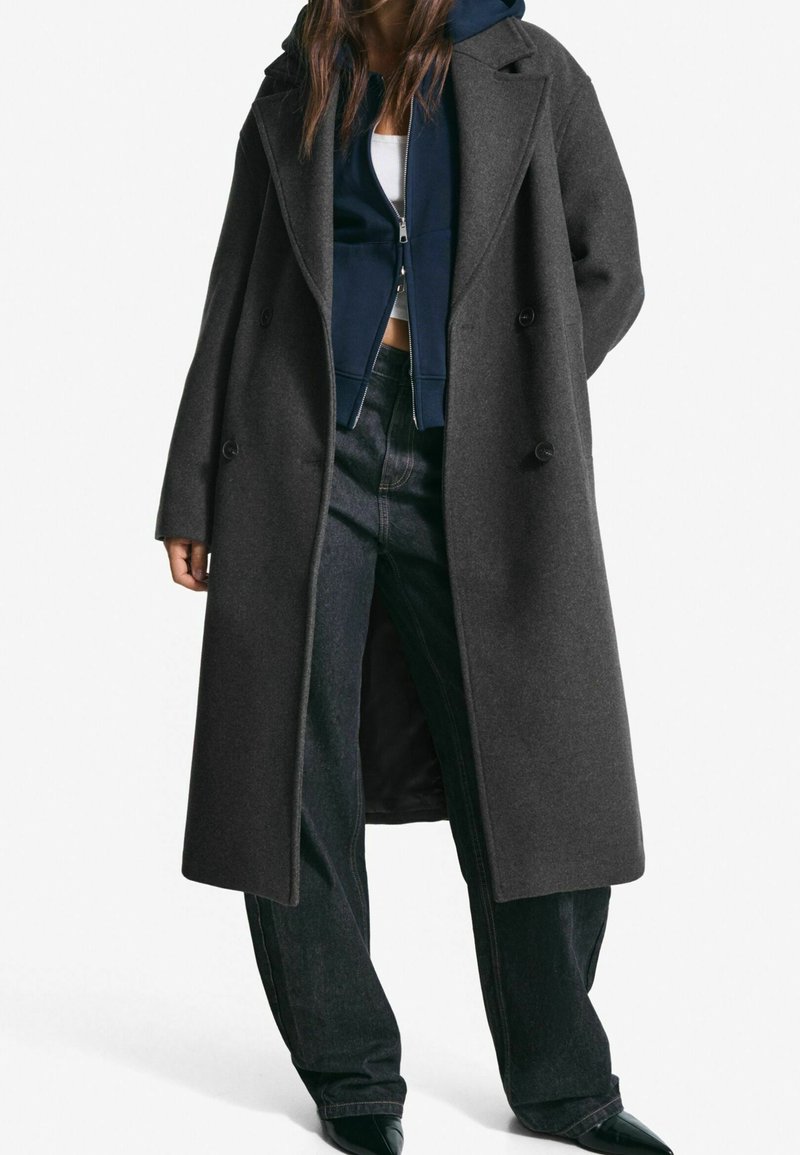 Femme portant un long manteau gris foncé à double boutonnage par-dessus un sweat à capuche bleu marine zippé, un crop top blanc, un jean noir ample et des chaussures noires pointues.