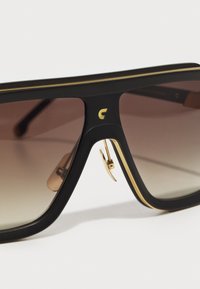 Gafas de sol negras y doradas con lentes marrones degradados, con un logo en relieve dorado en el puente y detalles dorados a lo largo de los bordes de la montura.