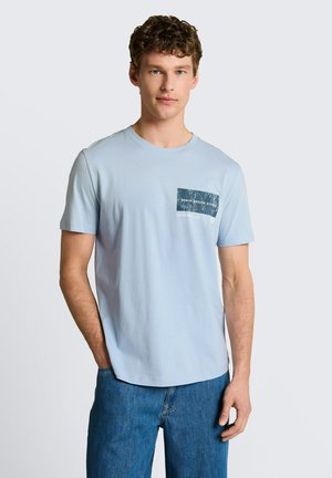 Młody mężczyzna z kręconymi włosami ubrany w jasnoniebieską koszulkę z ciemnoniebieskim wzorem kieszeni oraz niebieskie dżinsy, stoi na jednolitym tle.