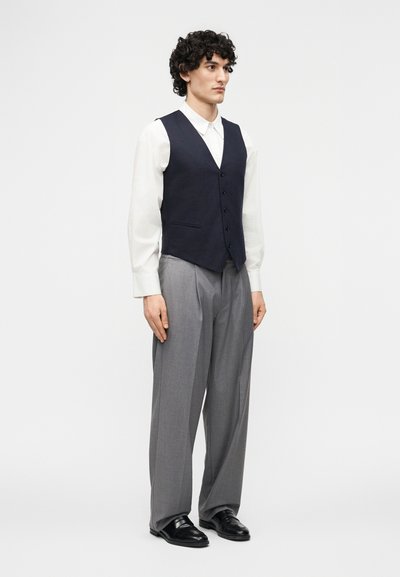 Άνδρας όρθιος, φοράει λευκό πουκάμισο μακρυμάνικο, σκούρο ναυτικό μπλε γιλέκο με κουμπιά, χαλαρό γκρι παντελόνι και μαύρα επίσημα παπούτσια σε απλό φόντο.