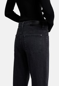 Jeans in denim nero con un design affusolato, dotati di due tasche posteriori e una texture sottile. Il cinturino presenta un'etichetta in pelle con logo.
