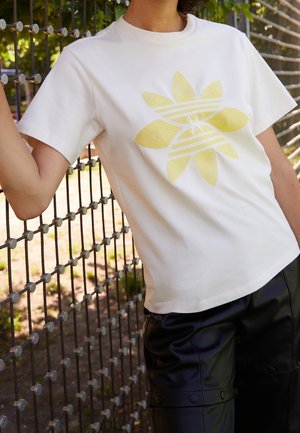 Osoba opierająca się o metalowy płot, ubrana w białą koszulkę z dużym żółtym geometrycznym wzorem kwiatowym oraz czarne skórzane spodnie na zewnątrz.