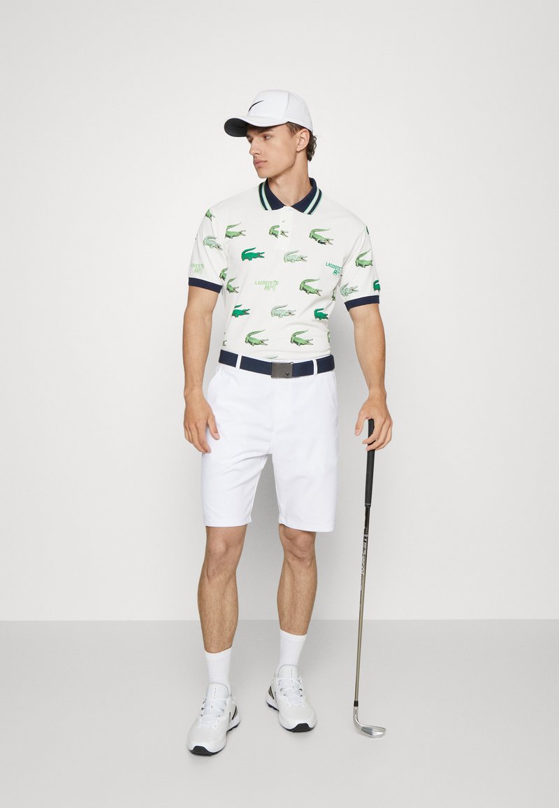 Chemise polo blanche avec des motifs de crocodile verts, col marine ; short blanc ; ceinture grise ; baskets blanches ; tenant un club de golf.