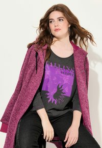 Cappotto viola strutturato sopra una maglietta a maniche lunghe grigio scuro con una stampa viola vivace. Jeans neri e grandi orecchini a cerchio completano l'outfit.