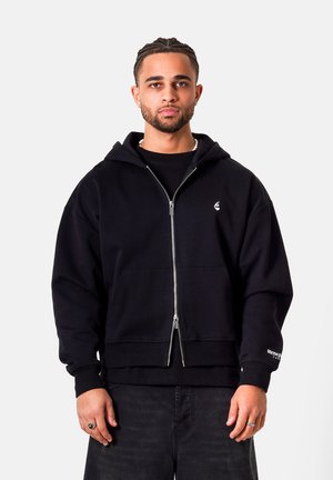 Młody mężczyzna z warkoczami, ubrany w czarną bluzę z zamkiem i ciemne dżinsy, stoi na jednolitym jasnym tle.