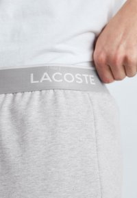 Szare sportowe szorty z elastycznym pasem, na którym znajduje się biała nazwa "LACOSTE". Materiał wygląda na miękki o gładkiej fakturze.