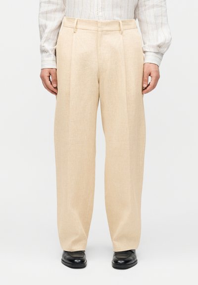 Personne portant un pantalon beige à jambes larges, une chemise blanche rayée à manches longues et des chaussures en cuir noires, debout sur un fond uni.
