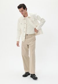 Jachetă din denim alb, cămașă albă cu nervuri, pantaloni kaki și pantofi negri. Modelul poziționat în mișcare, pe un fundal simplu.