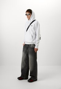 Witte hoodie met een voorzak, losse grijze denim jeans, zwart-rode schoenen en ronde zonnebril. Ketenaccent op de jeans.
