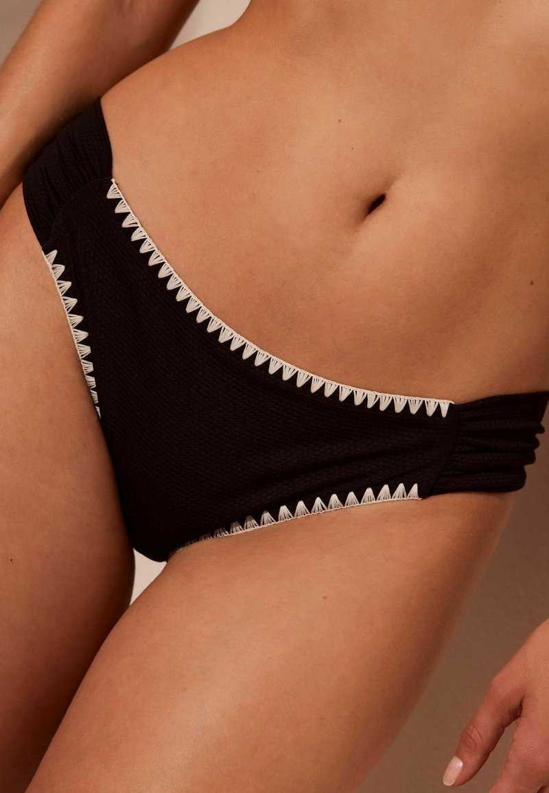 Fekete bikini alsó textúrált anyagból, fehér cikk-cakk díszítéssel a széleken, és ráncolt oldali részletekkel a kényelmes illeszkedés érdekében.