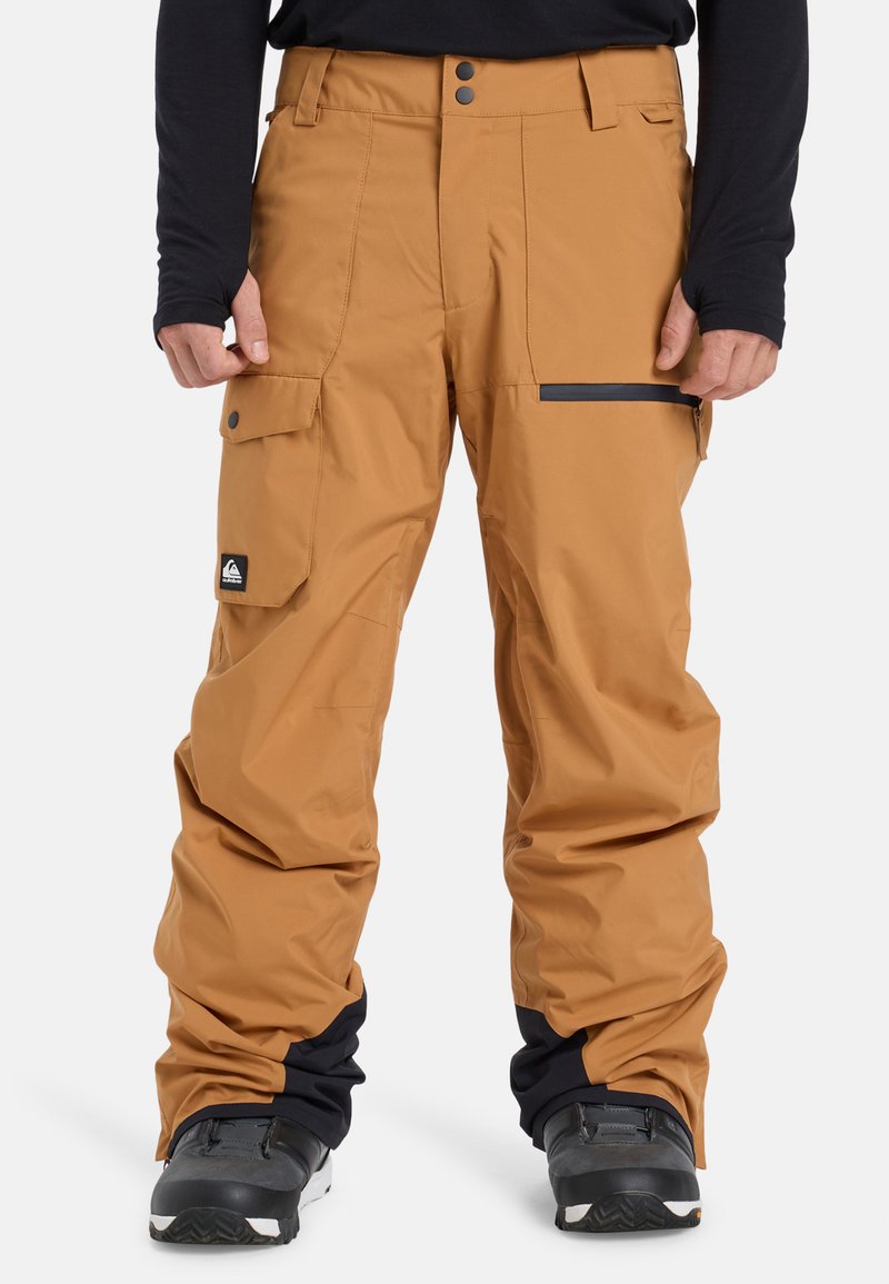 Braune Schneehose mit lockerer Passform, ausgestattet mit Seitentaschen, einer schwarzen Reißverschlusstasche und einem Logopatch nahe dem Saum. Wasserabweisendes Material.